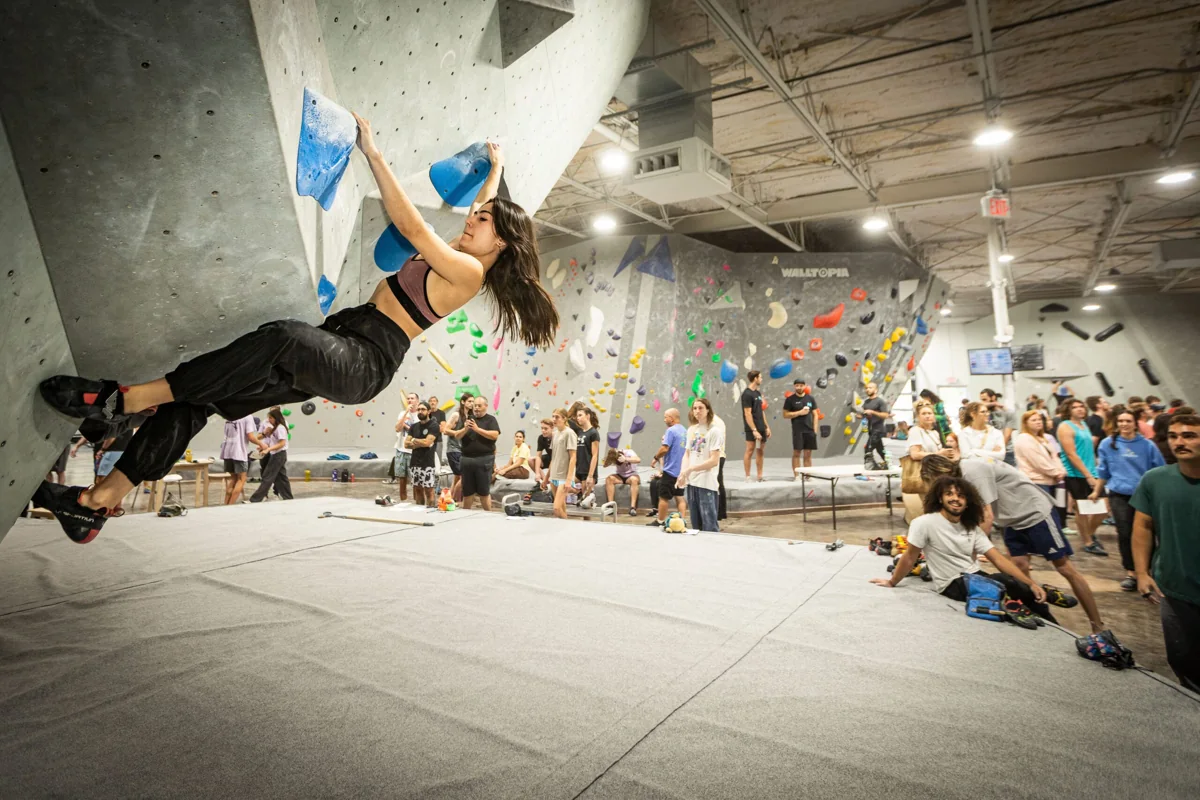 A climber moves across an angular wall while a crowd watches inside Blue Swan Boulders.